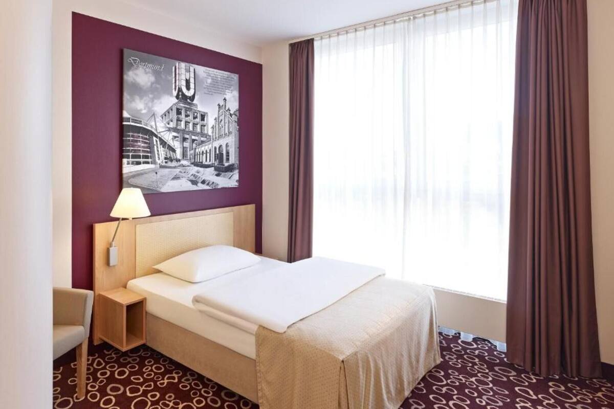 A hotel room with a bed, a nightstand, a lamp, a window with curtains, and a wall with a cityscape artwork.