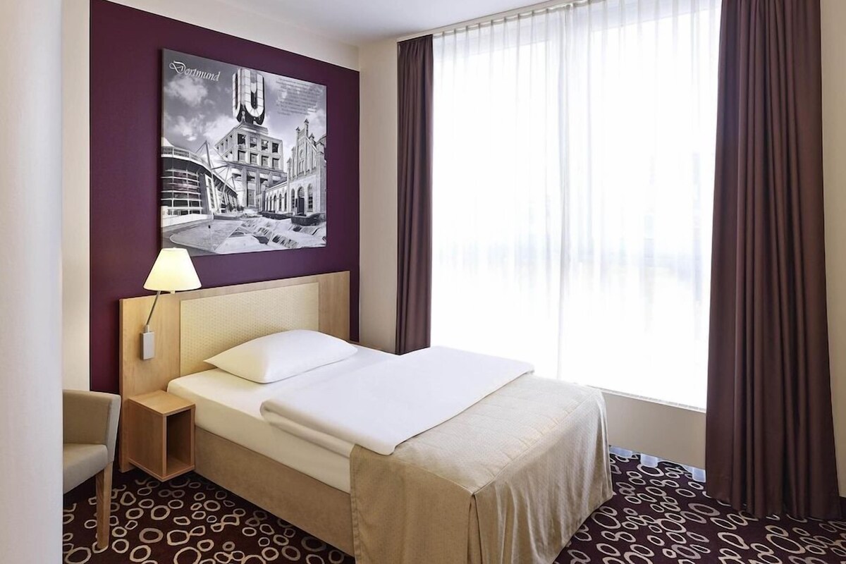 A hotel room with a bed, a nightstand, a lamp, a window with curtains, and a wall-mounted picture.