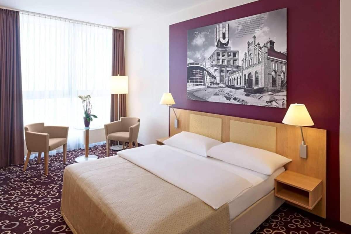 A hotel room with a bed, two chairs, a table with a plant, and a large black and white cityscape painting on the wall.