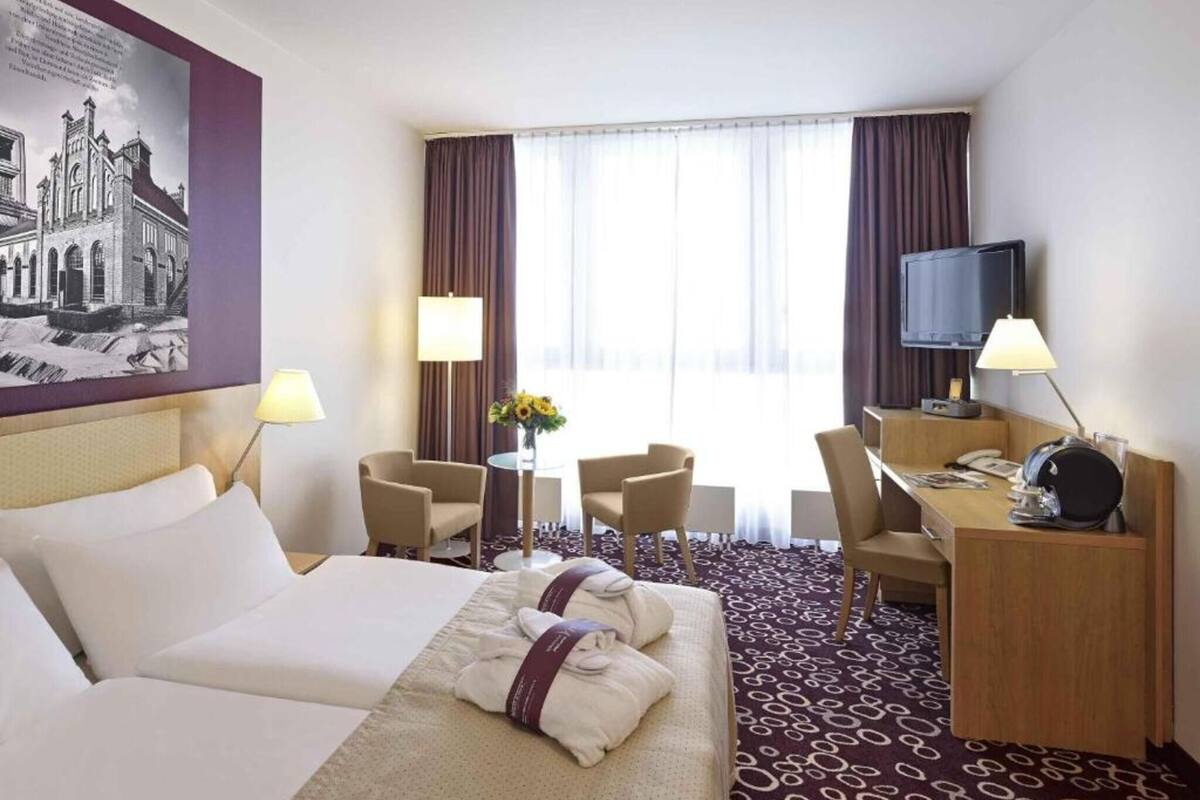 A hotel room with a bed, two chairs, a desk with a television, and a painting on the wall.