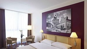 Habitación de hotel con una cama, dos sillas, una mesa con una planta y un cuadro grande en la pared.