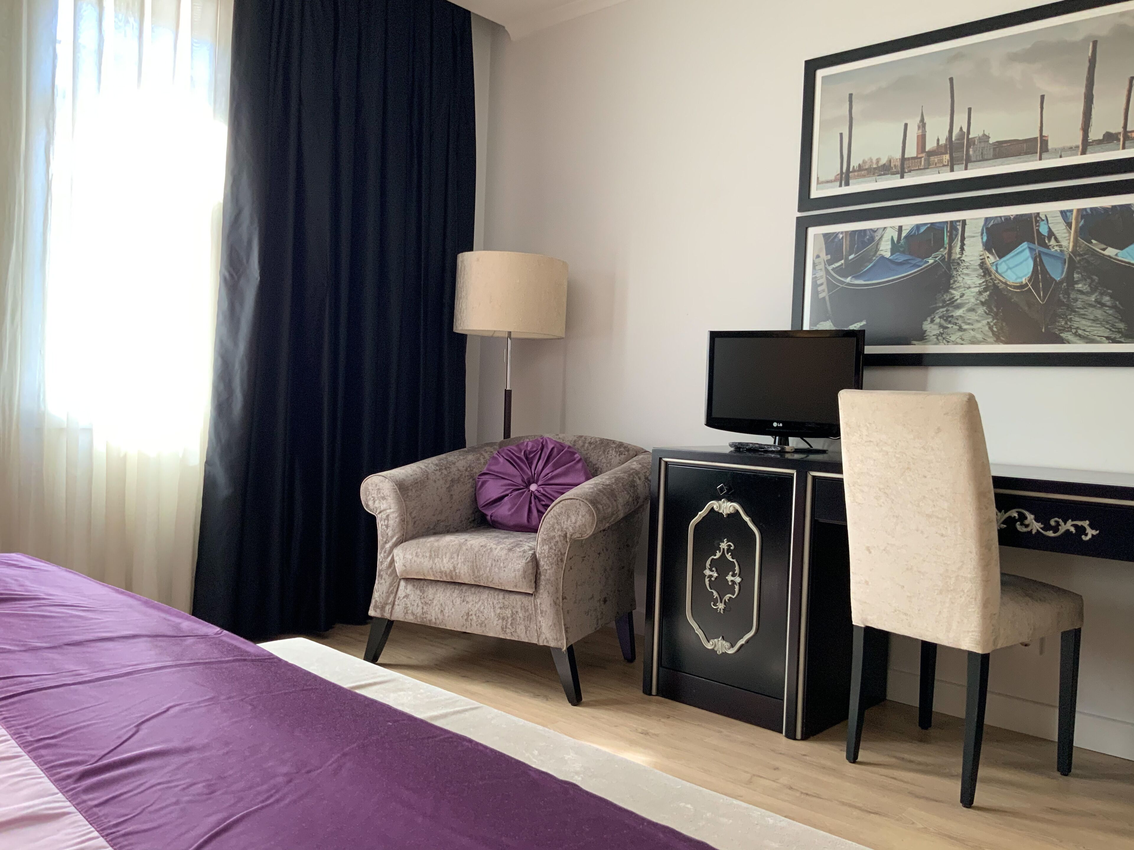 A hotel room with a bed, a desk with a computer, a chair, a lamp, and a large window with curtains.