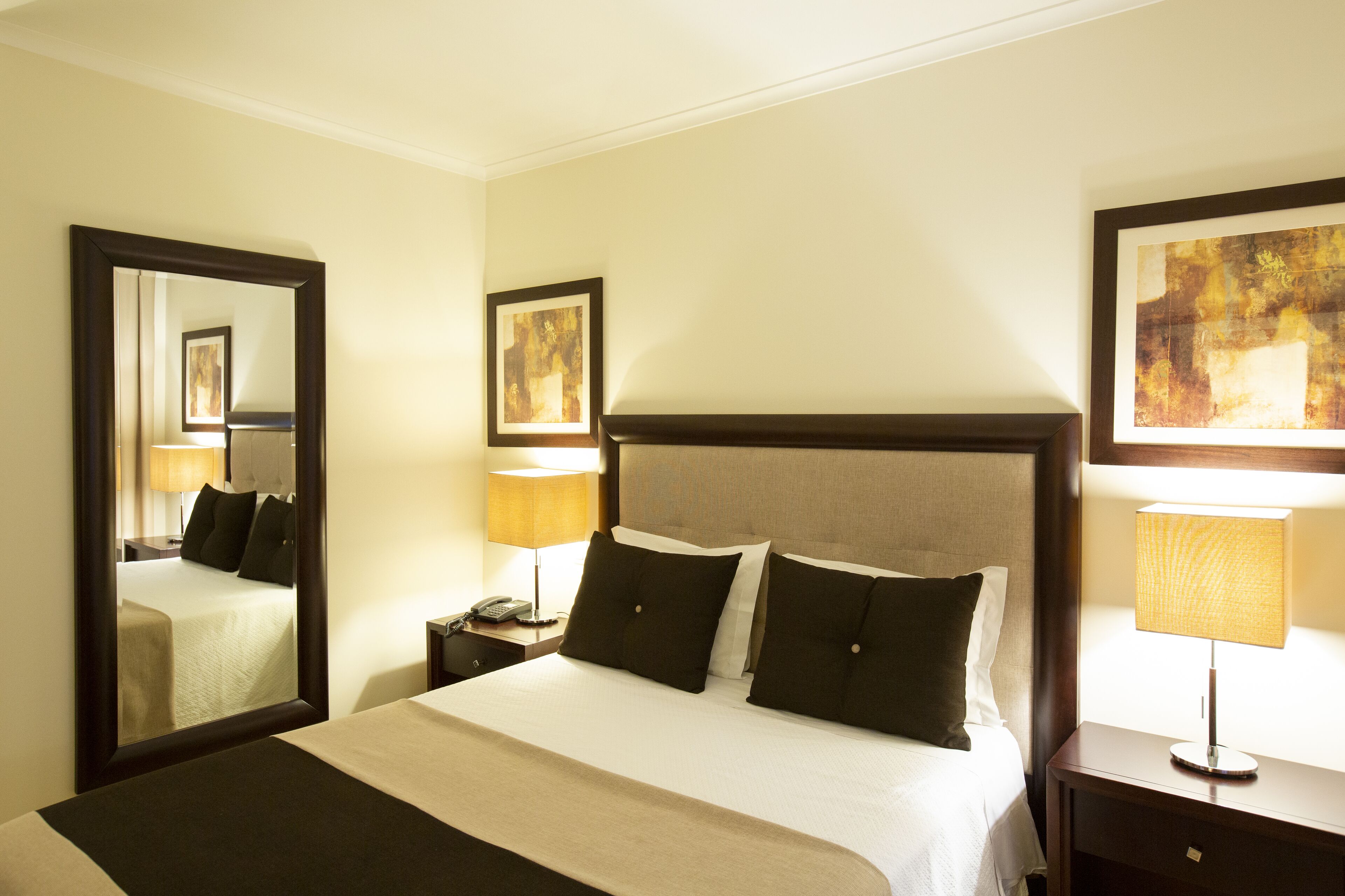 A hotel room with a large bed, two bedside tables, a nightstand, a mirror, and two framed artworks on the walls.