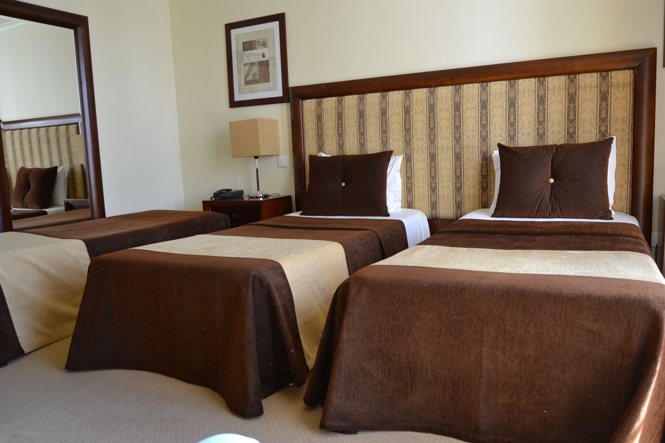 Habitación de hotel con tres camas individuales, cada una con cubrecama marrón y almohadas blancas. Se aprecia un cabecero de madera con diseño a rayas, una mesita de noche con lámpara y un cuadro enmarcado en la pared.