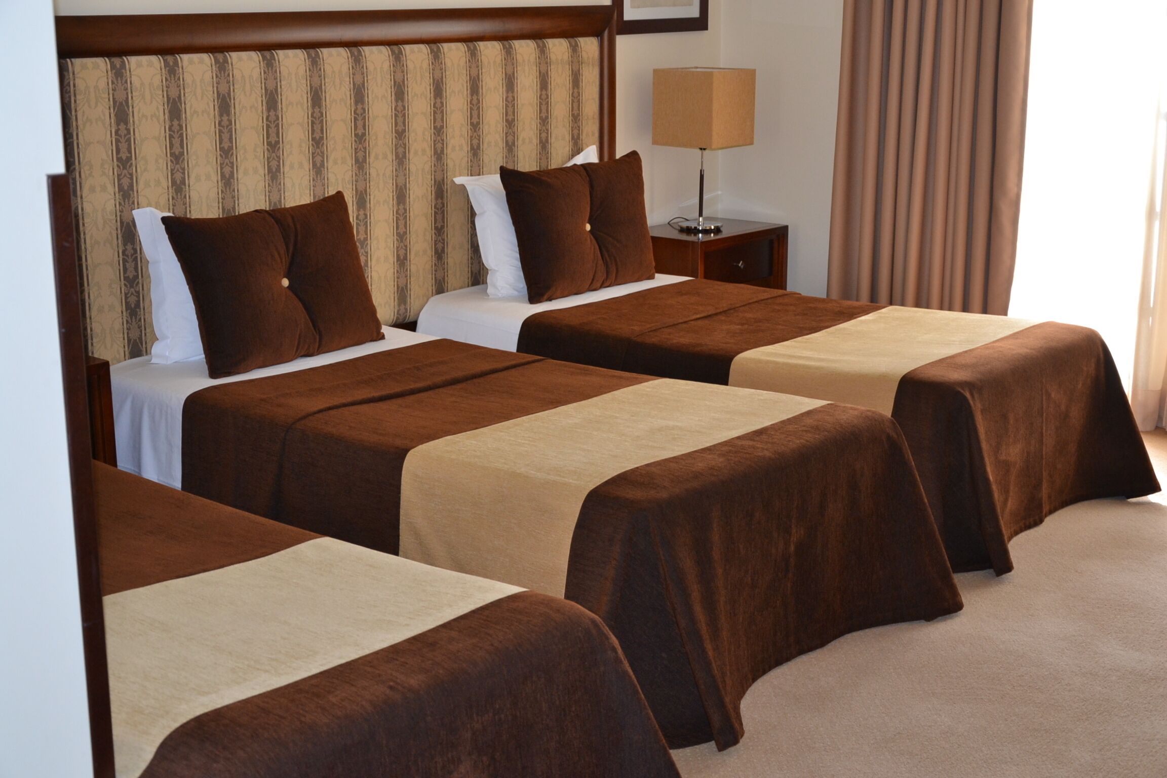 Habitación de hotel con tres camas individuales, cada una con cubrecama marrón y almohadas blancas. Se observa una mesita de noche con una lámpara y una pequeña mesa con una lámpara junto a las camas.