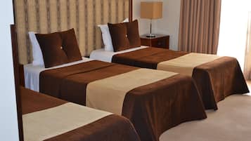 Une chambre d’hôtel avec trois lits simples, chacun recouvert d’une couverture marron et agrémenté d’oreillers blancs. On y trouve une table de chevet avec une lampe, ainsi qu’une petite table avec une lampe placée à côté des lits.
