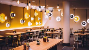 Un restaurante con varias mesas y sillas, una pared amarilla con lámparas circulares y bombillas colgantes.