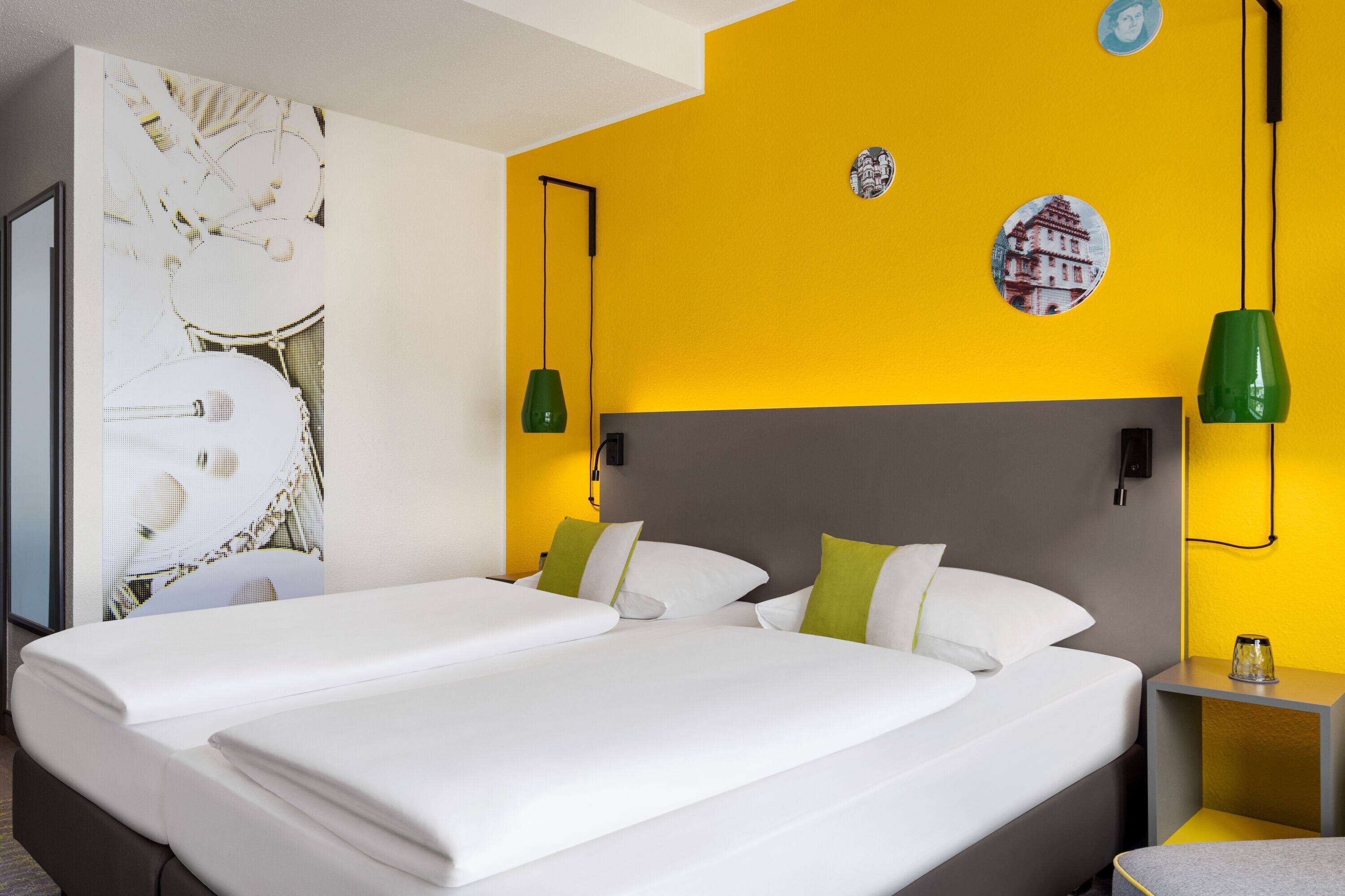 Cama doble con sábanas blancas, almohadas verdes y cabecera gris. Dos lámparas verdes fijadas a la pared y una mesita con un vaso encima.