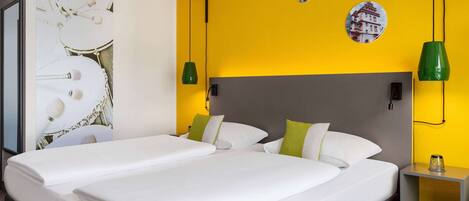 Cama doble con sábanas blancas, almohadas verdes y cabecera gris. Dos lámparas verdes fijadas a la pared y una mesita con un vaso encima.