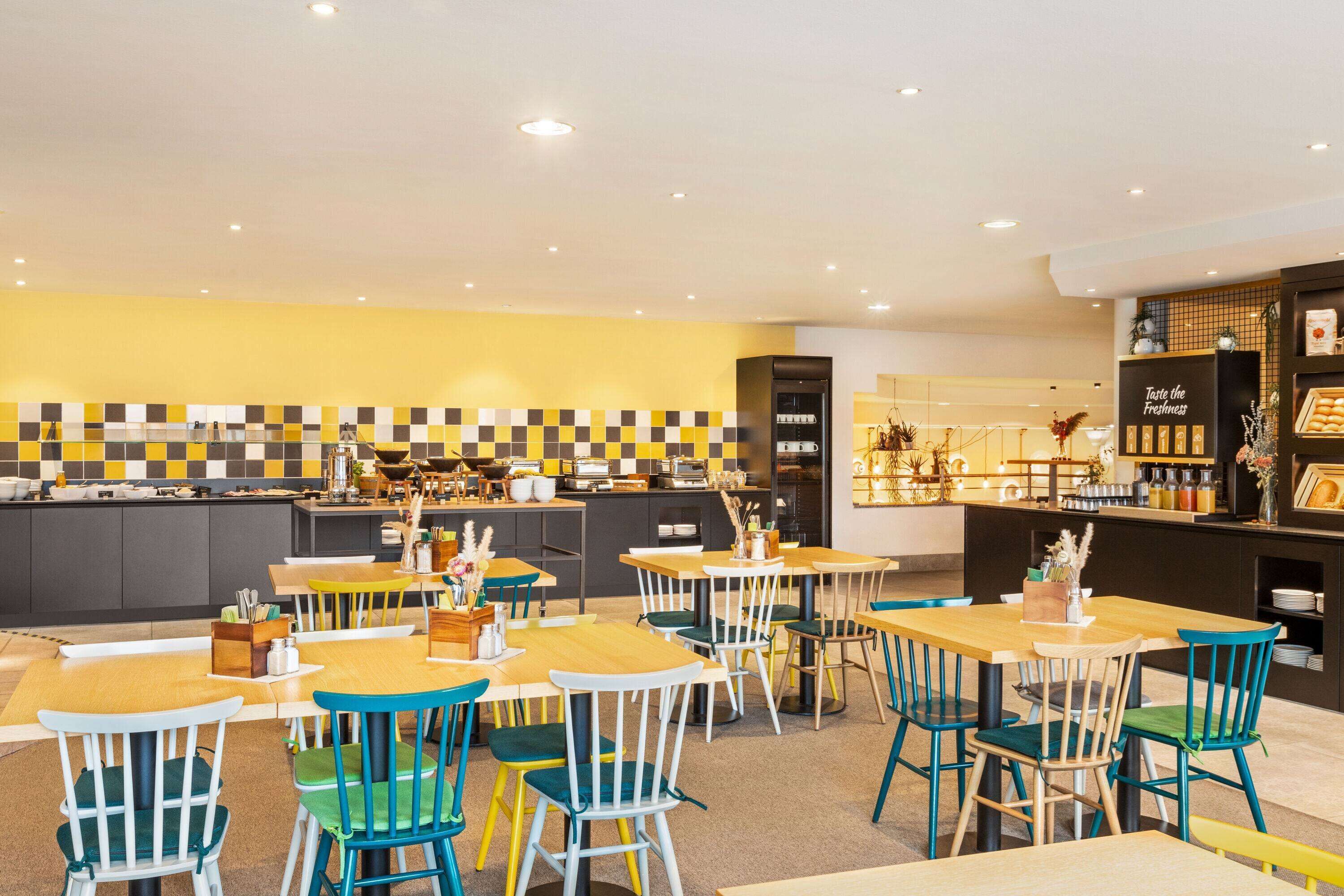 Un restaurante moderno con un salpicadero amarillo y negro a cuadros, mesas de madera y sillas de colores.