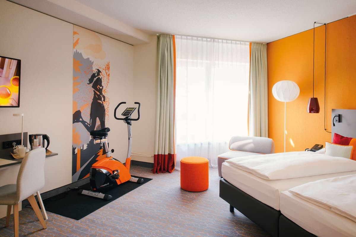 A hotel room with a bed, a desk, a chair, an exercise bike, and a large window with curtains.