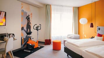 Habitación de hotel con cama, escritorio, silla, bicicleta estática y un amplio ventanal con cortinas.