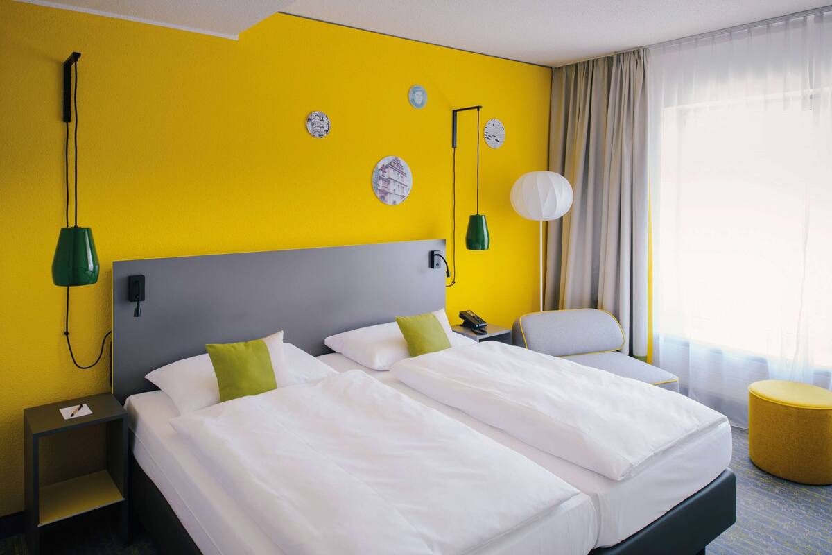 A hotel room with a double bed, a bedside table, a lamp, a chair, and a window with curtains.