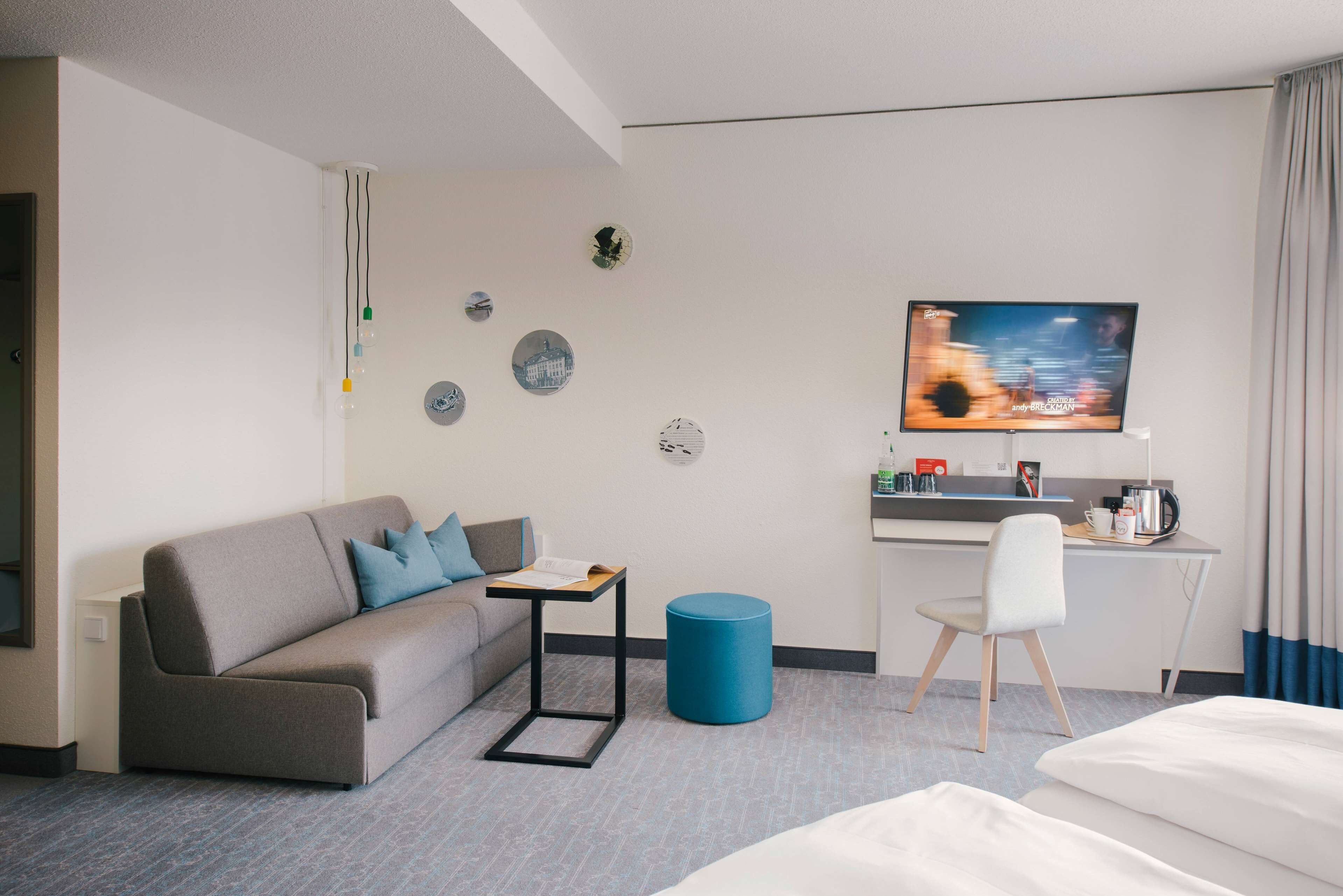 A hotel room with a grey sofa, a small table, a blue ottoman, a white chair, a bed, a desk with a TV, and a coffee machine.