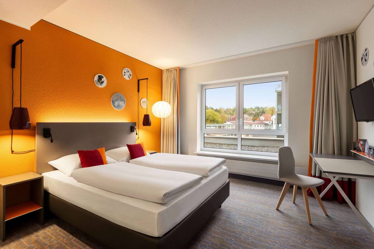 A hotel room with a bed, a desk, a chair, and a window with a view of buildings.