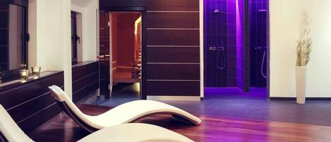 Una moderna stanza spa con pavimento in legno, una poltrona relax, un tavolino con candele e una zona doccia illuminata di viola.