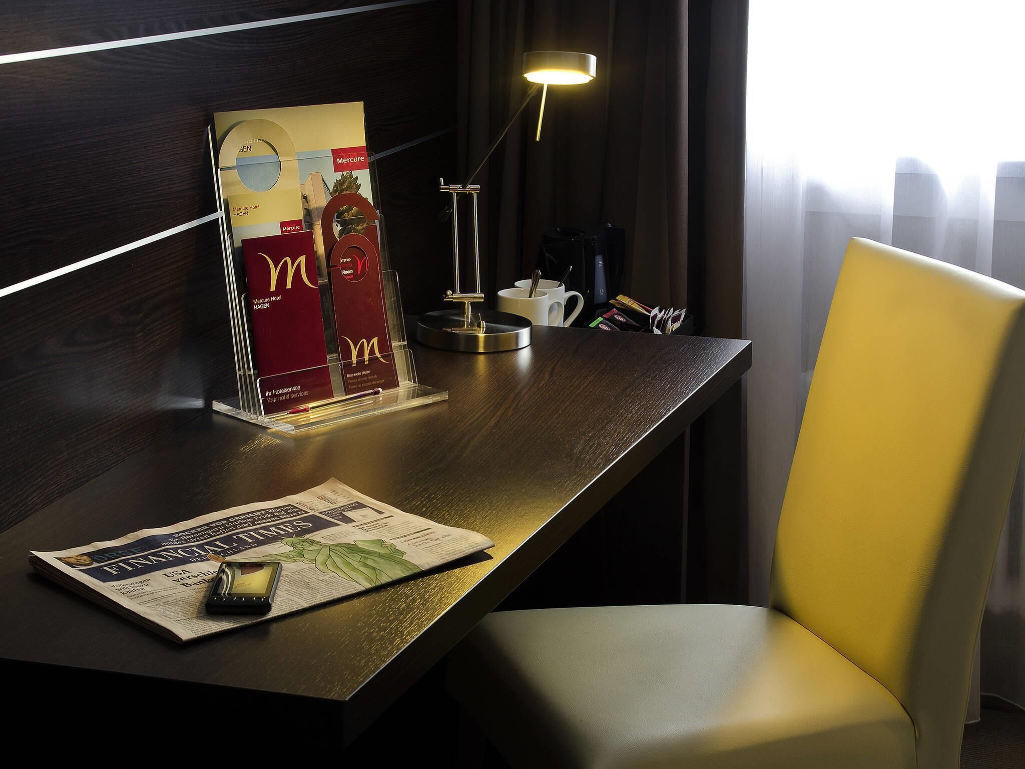 A hotel desk with a newspaper, a mobile phone, a lamp, and a chair.