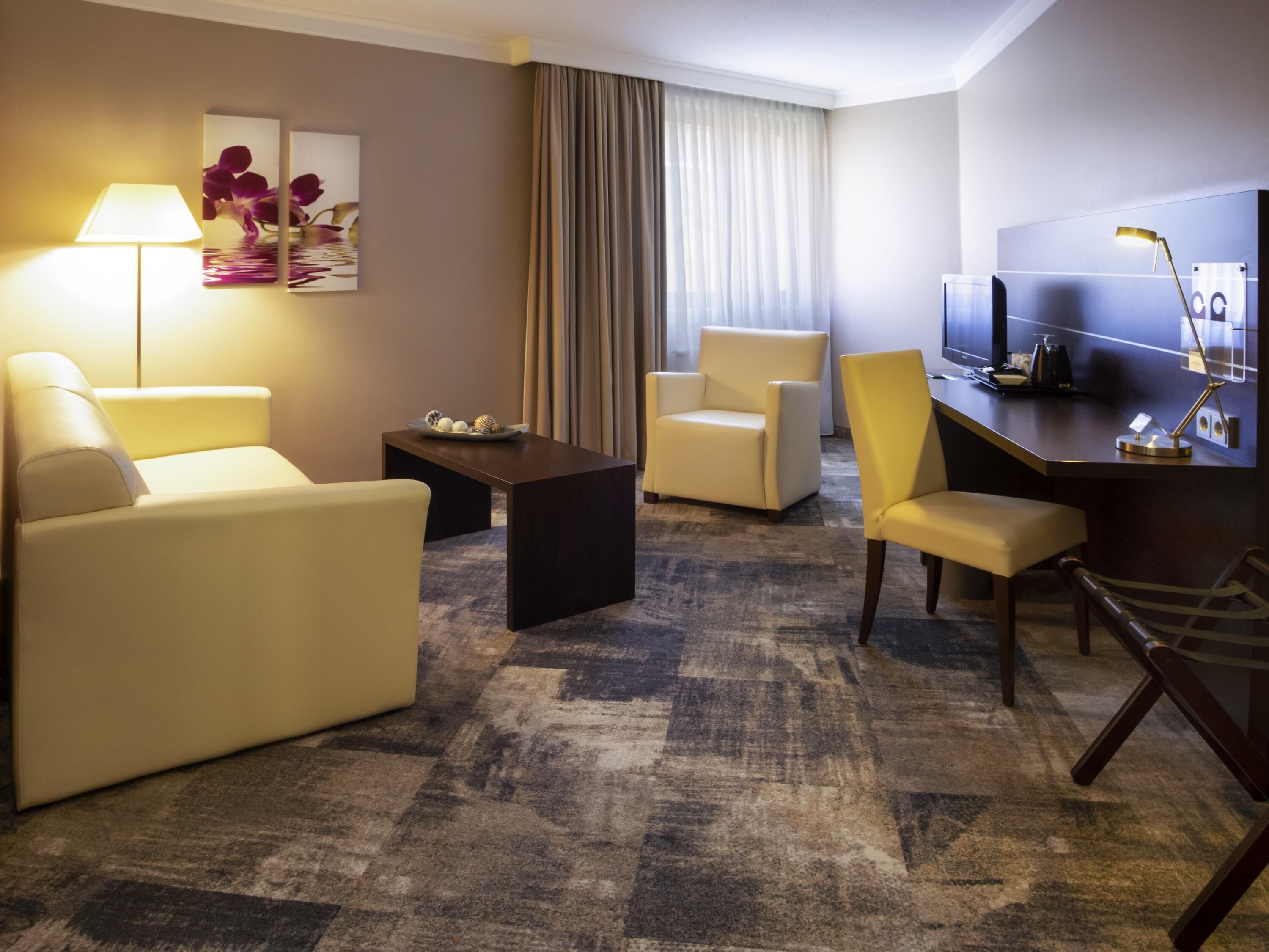 A hotel room with a sofa, armchair, desk, television, lamp, and artwork.