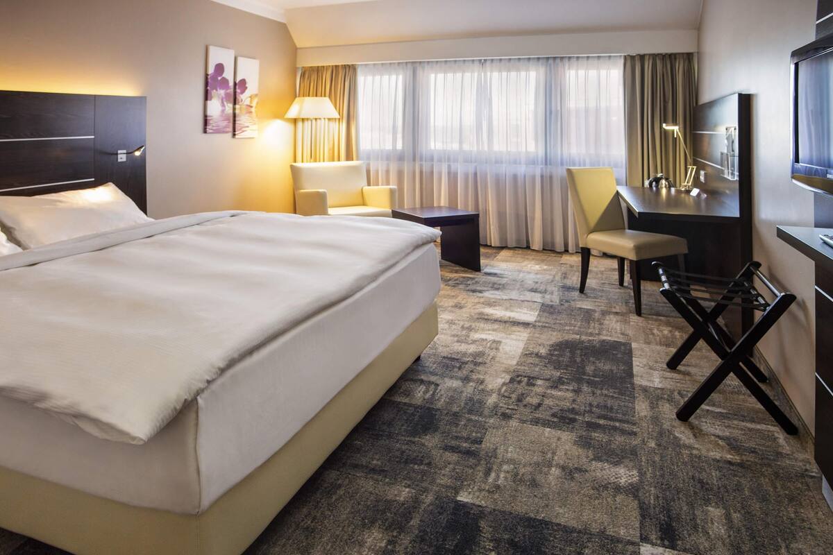 A hotel room with a large bed, a desk, a chair, a TV, and a window with curtains.