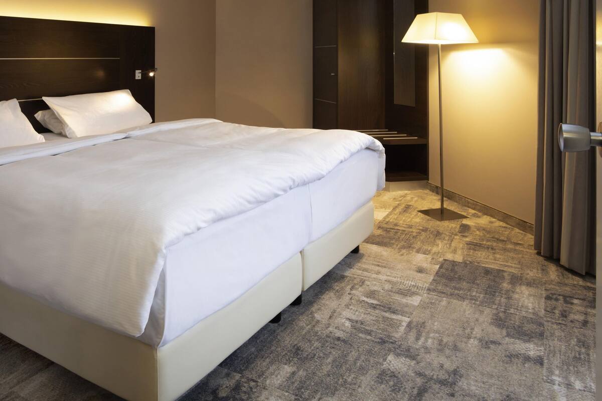 A modern hotel room with a large bed, a nightstand, a lamp, and a wardrobe.