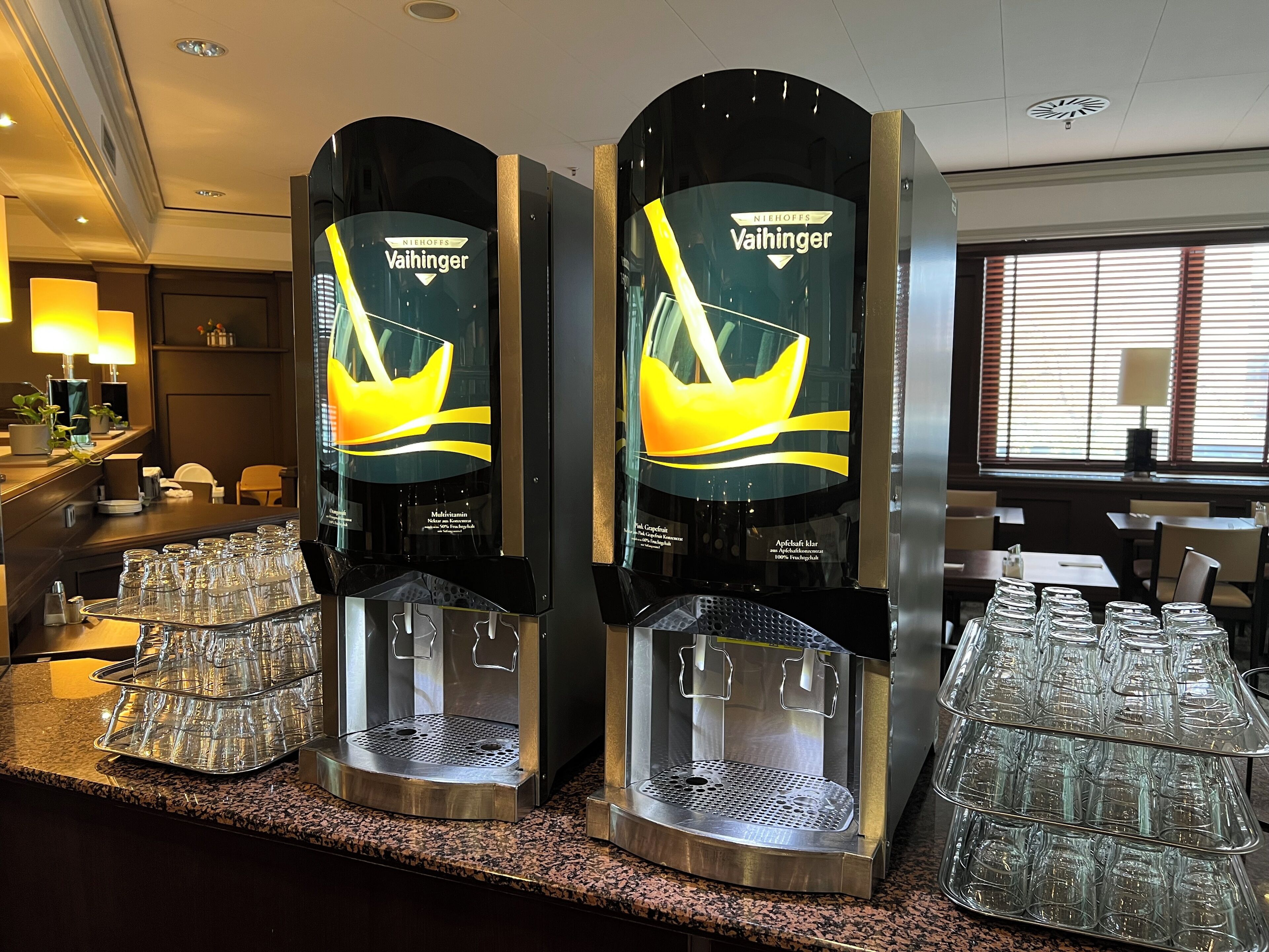 A bar area with two beverage dispensers, a countertop with glassware, and a window with blinds.