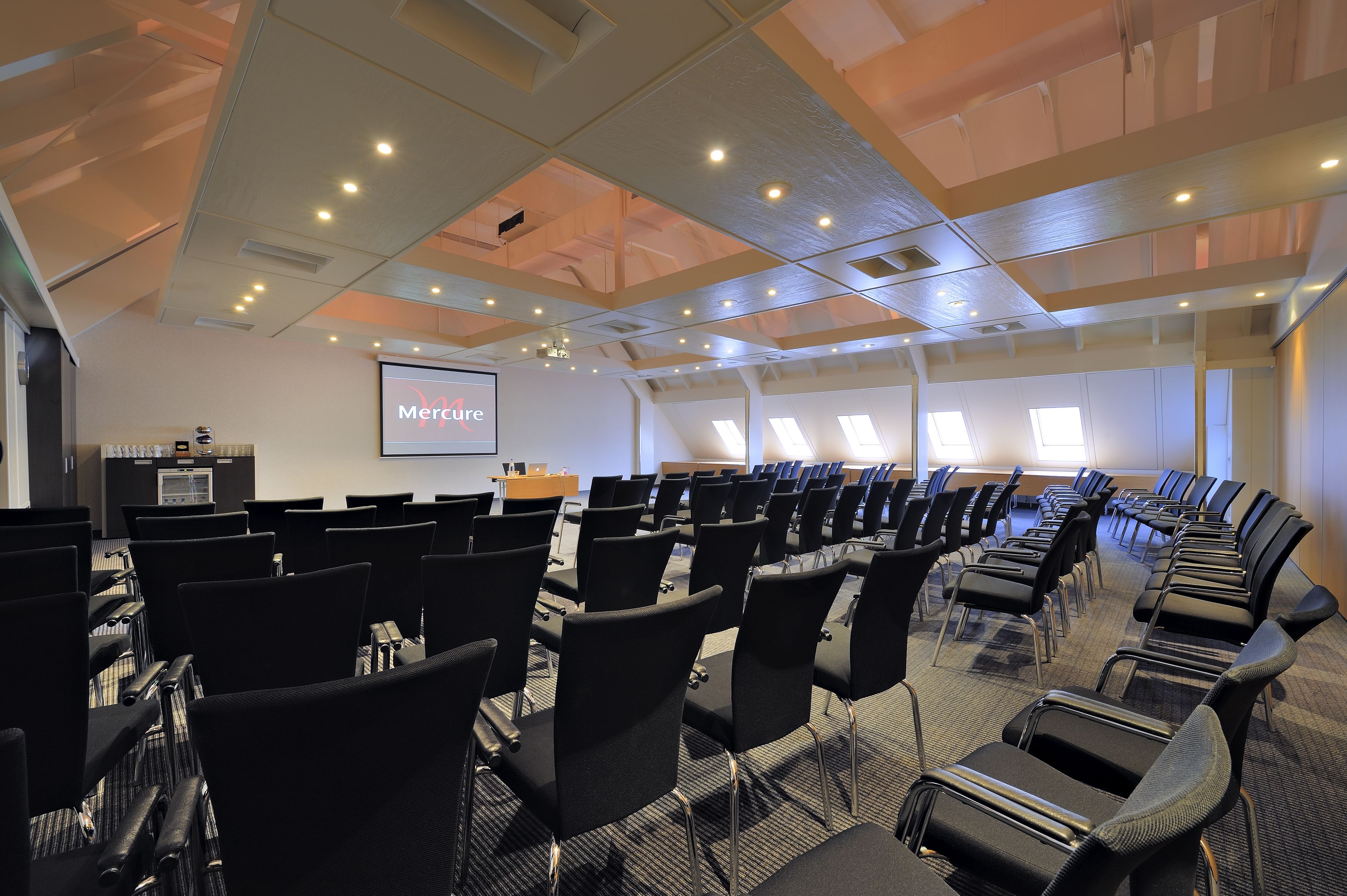 A conference room with rows of black chairs, a projector screen displaying 'Mature', and a podium with a laptop.