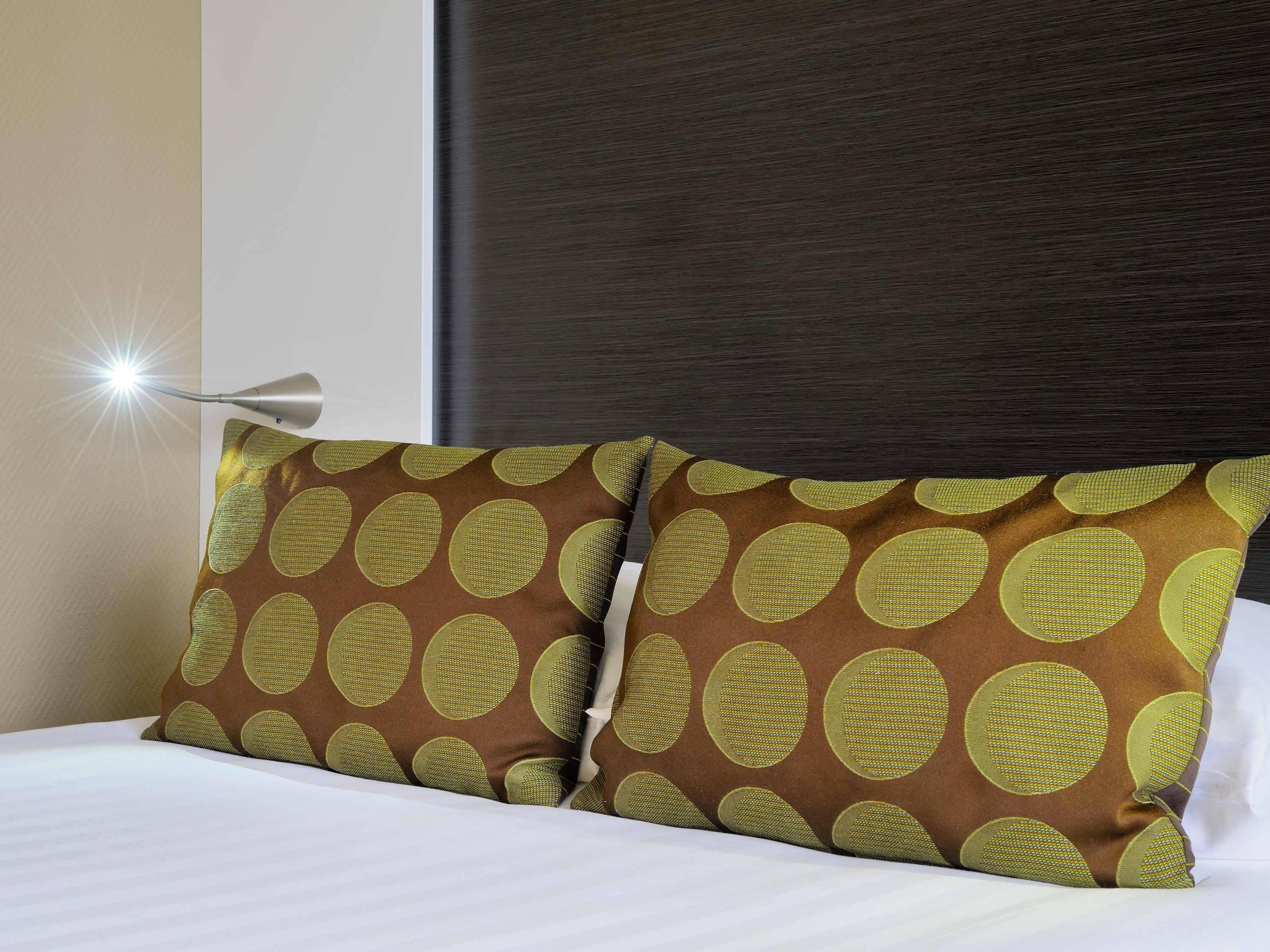 Una cama bien hecha con dos almohadas de diseño circular, una lámpara de noche y un cabecero oscuro.