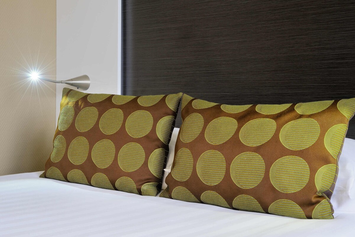 Een keurig opgemaakt bed met twee kussens met een rond patroon, een bedlampje en een donker hoofdbord.