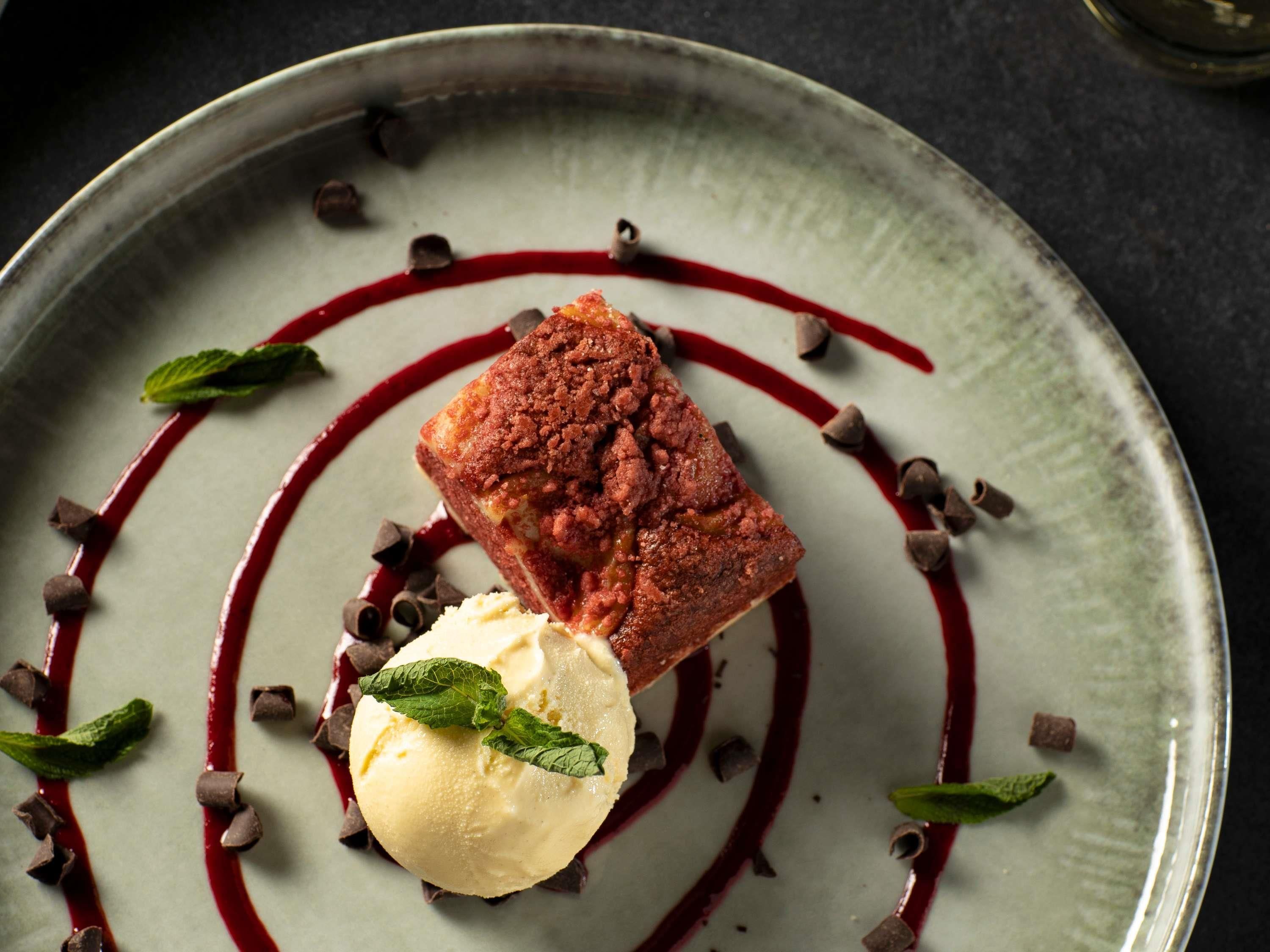 Um prato de sobremesa com um pedaço quadrado de bolo, uma bola de sorvete, raspas de chocolate e um molho vermelho.
