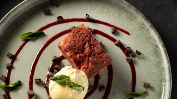 A dessert plate with a square piece of cake, a scoop of ice cream, chocolate shavings, and a red sauce.