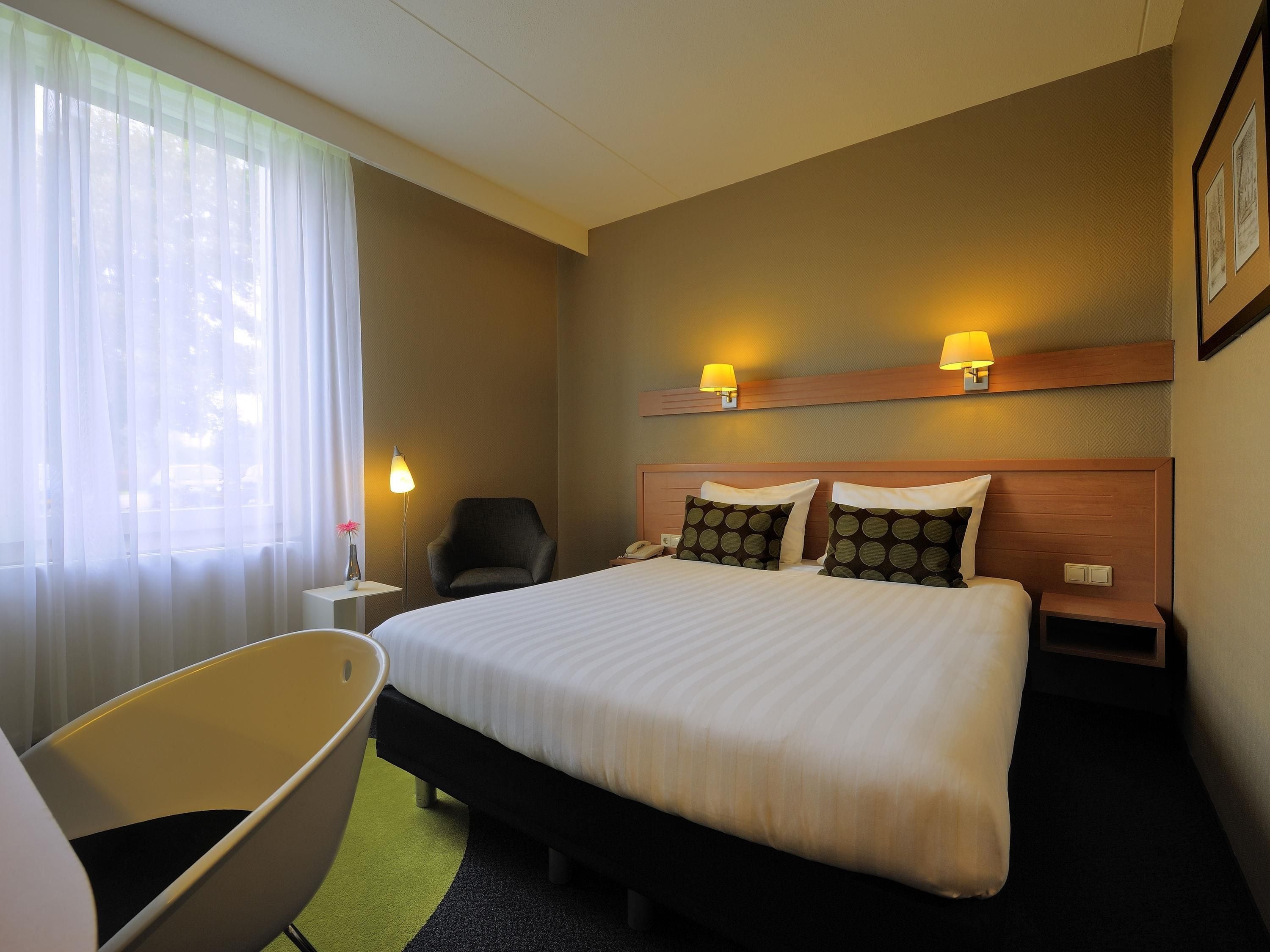 Een hotelkamer met een groot bed, twee bedlampen, een stoel en een kleine tafel met een lamp.