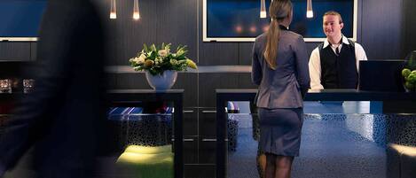 Un espace d’accueil moderne comprenant un comptoir de réception, un vase de fleurs et un coin salon avec un pouf vert.