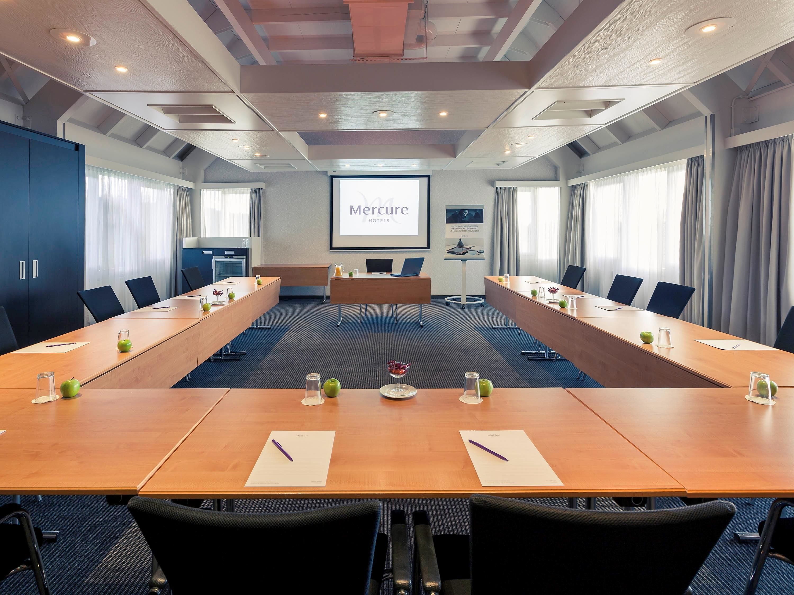 A conference room with a long table, chairs, a projector screen, and a presentation about Mercure Hotels.