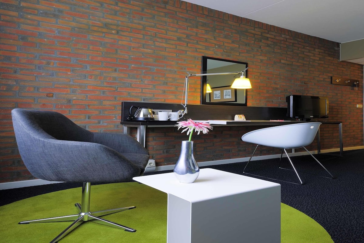 Een hotelkamer met een bakstenen muur, een bureau met een flatscreen televisie, een moderne stoel, een klein tafeltje met een vaas, en een groen tapijt.