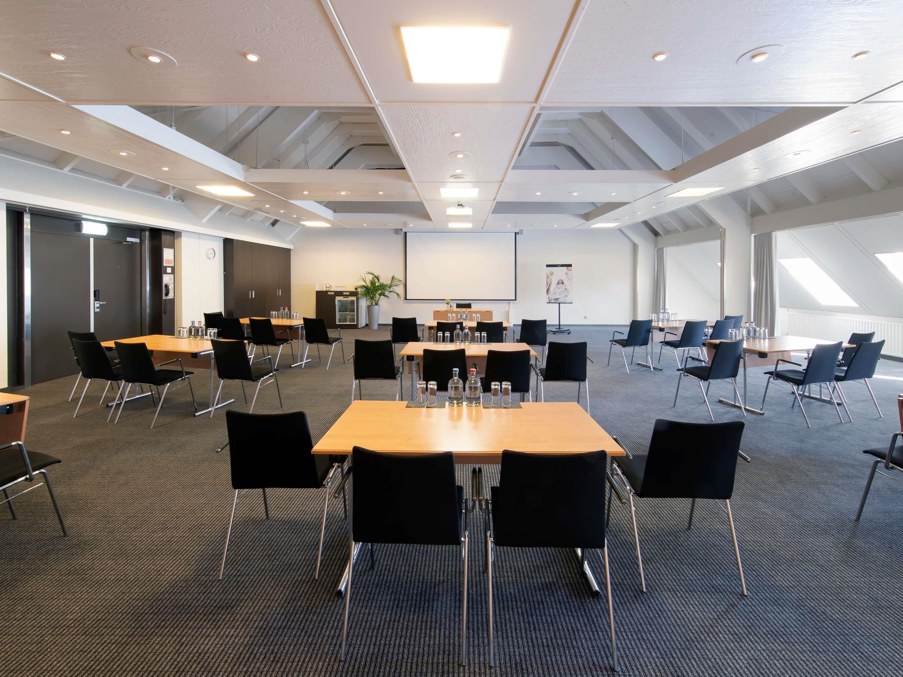 A conference room with multiple tables, chairs, a projector screen, and a large window.