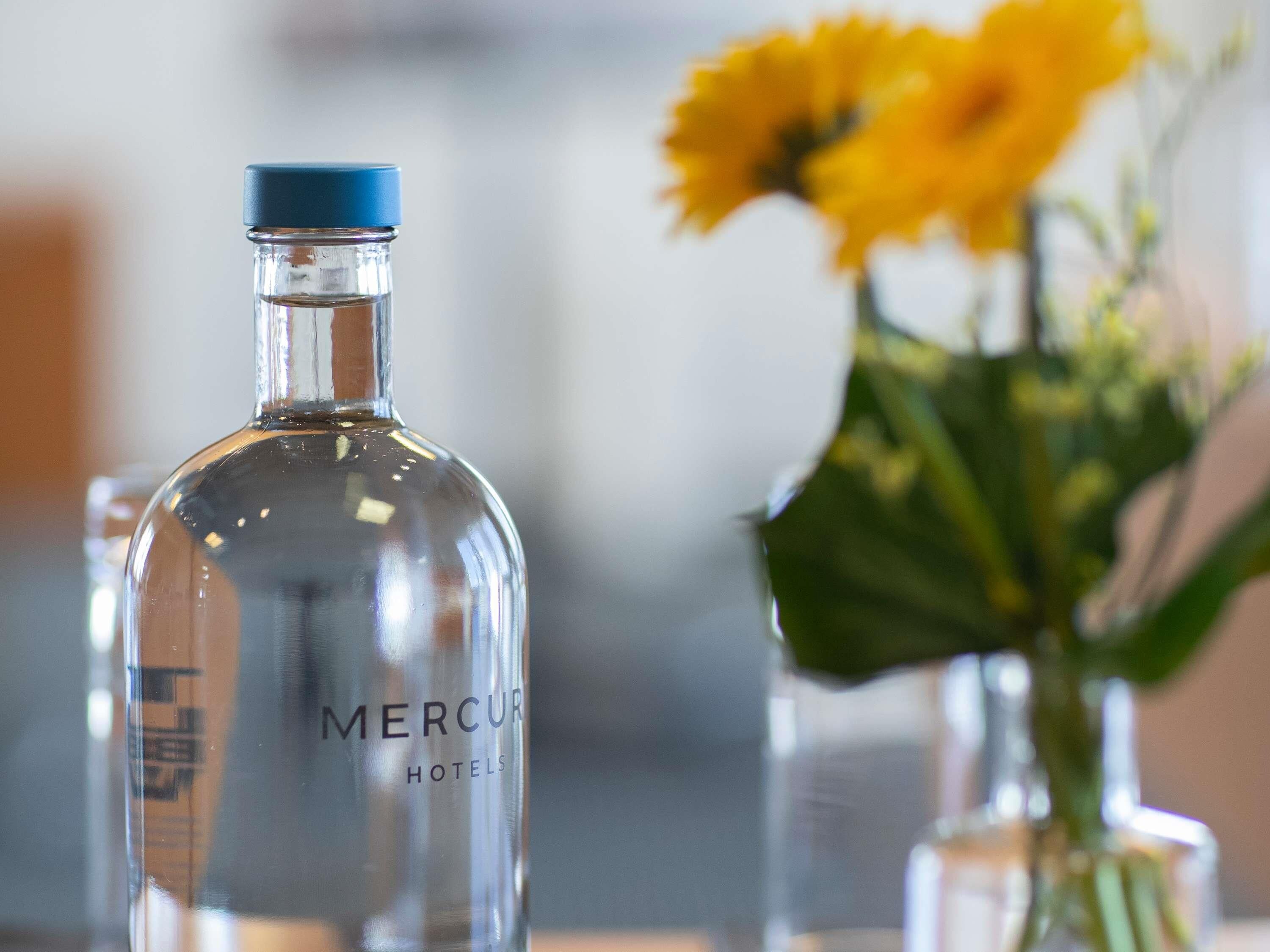 Una botella de vidrio transparente con la etiqueta "MERCUR HOTELS" y un jarrón con flores amarillas al fondo.