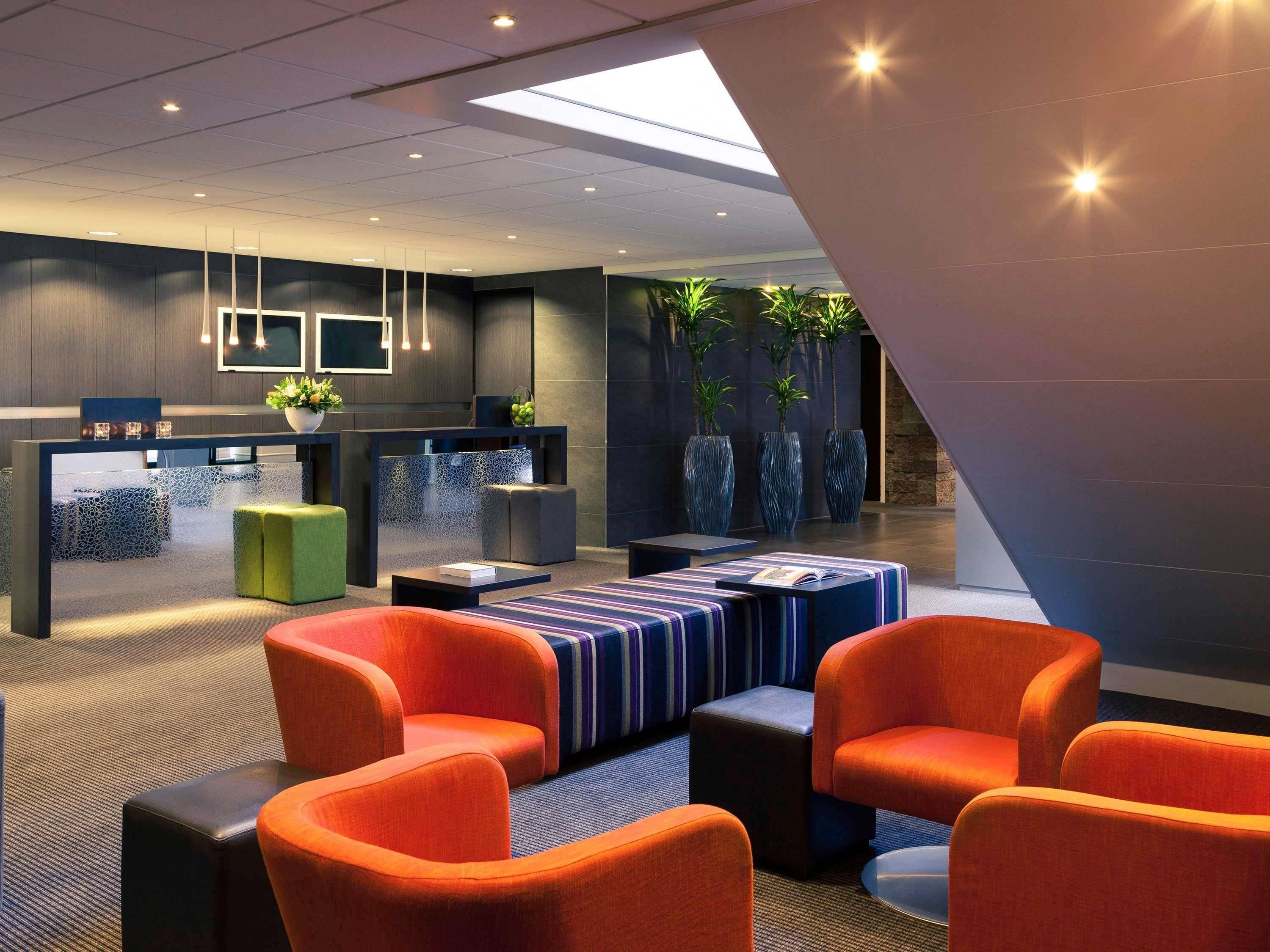 Eine moderne Lobby mit einer Rezeption, einer Sitzgruppe mit orangefarbenen Sesseln und Zierpflanzen.