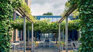 An outdoor seating area with tables and chairs under a pergola covered with vines, surrounded by brick buildings. - Mercure City Zwolle Hotel (Zwolle)
