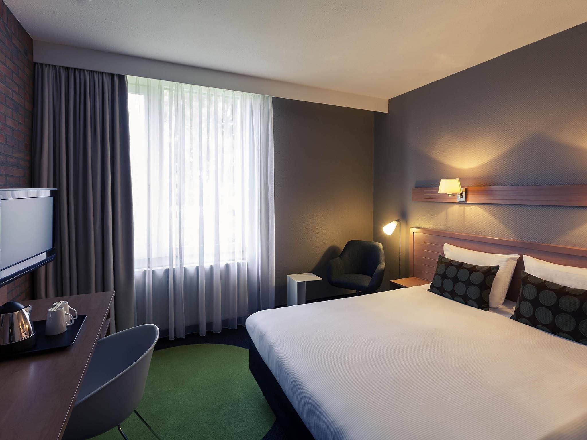A hotel room with a bed, a desk with a chair, a TV, a lamp, and a window with curtains.
