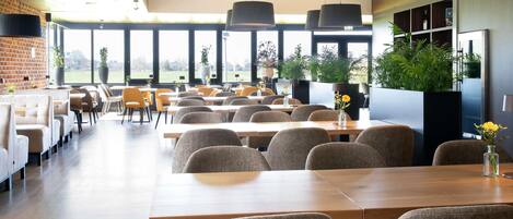 A spacious dining area with long tables and chairs, large windows with a view of greenery, and modern pendant lighting.