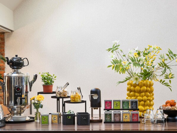 A coffee station with a coffee machine, a teapot, a potted plant, a jar of pickles, a small table with tea bags, and a vase with flowers.