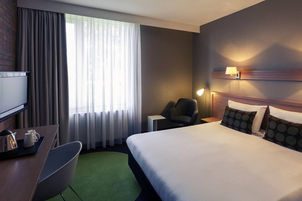 Une chambre d’hôtel comprenant un lit, un bureau avec une chaise, une télévision, une lampe et une fenêtre avec des rideaux.