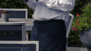 Eine Person hält ein Tablett mit einer Flasche und zwei Gläsern, vermutlich um Wein zu servieren, in einer Außenanlage mit Grünpflanzen im Hintergrund.