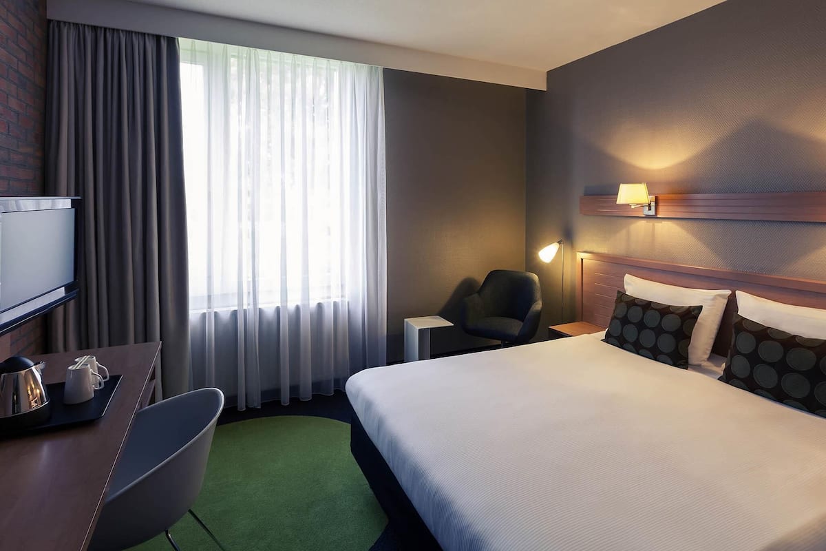 Een hotelkamer met een bed, een bureau met stoel, een televisie, een lamp en een raam met gordijnen.