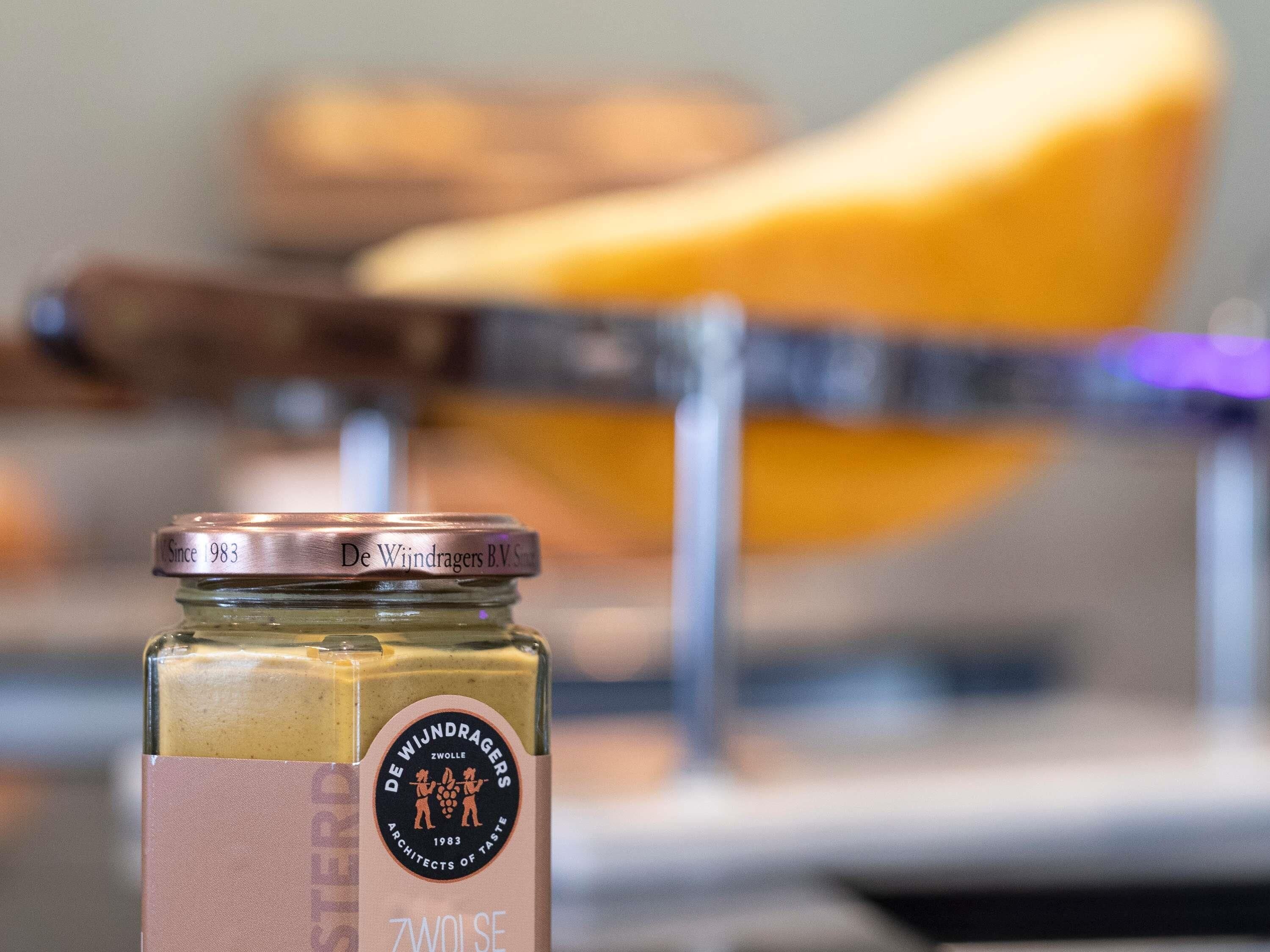 Un frasco de queso ZWOLSE con tapa metálica, colocado frente a un cortador de queso.