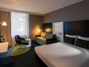 Una habitación de hotel con cama, escritorio, silla, lámpara y ventana con cortinas.