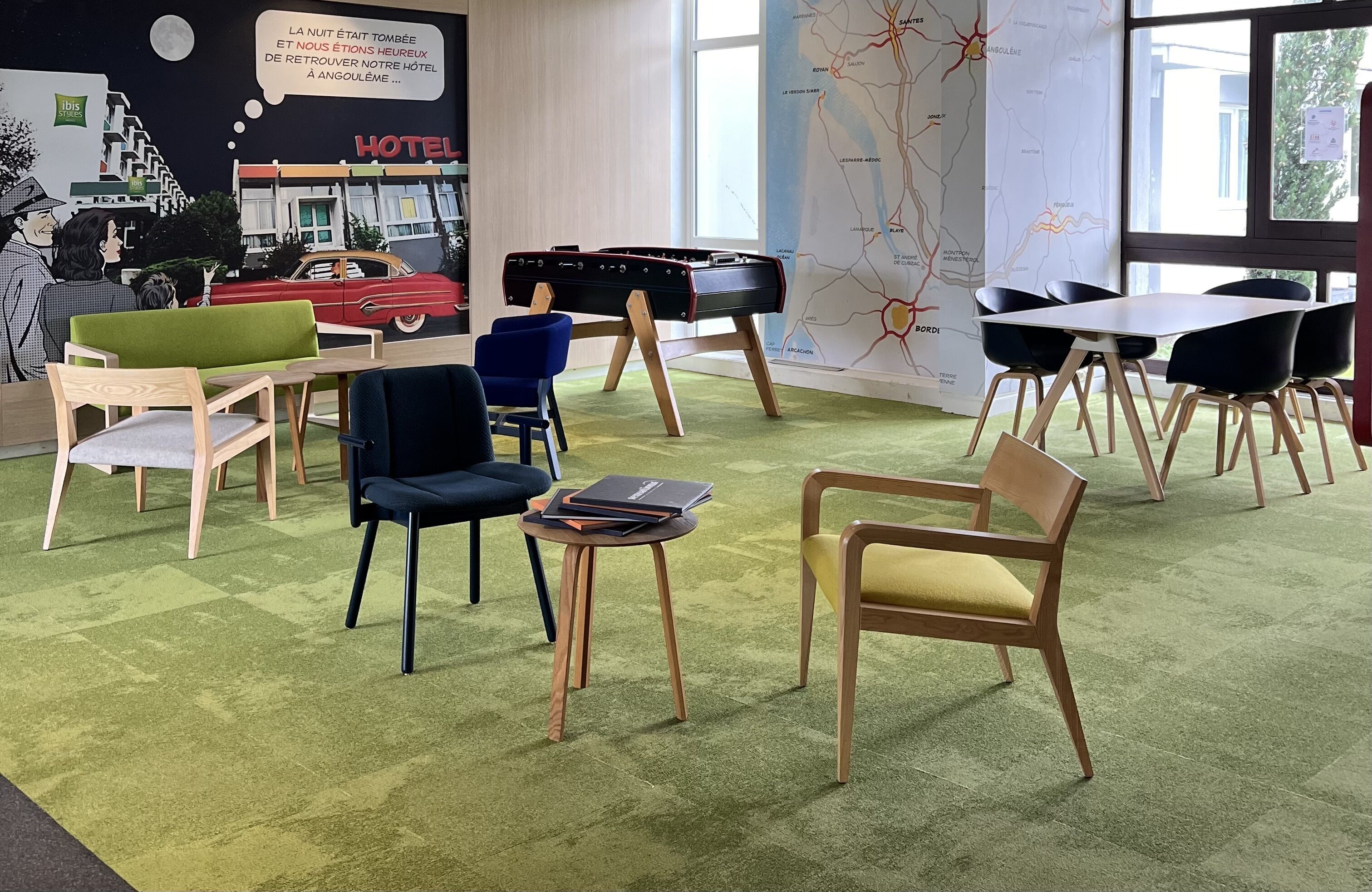 Un vestíbulo de hotel con alfombra verde, sillas de madera, una silla azul, una mesita con un libro y un mural que representa una escena hotelera.