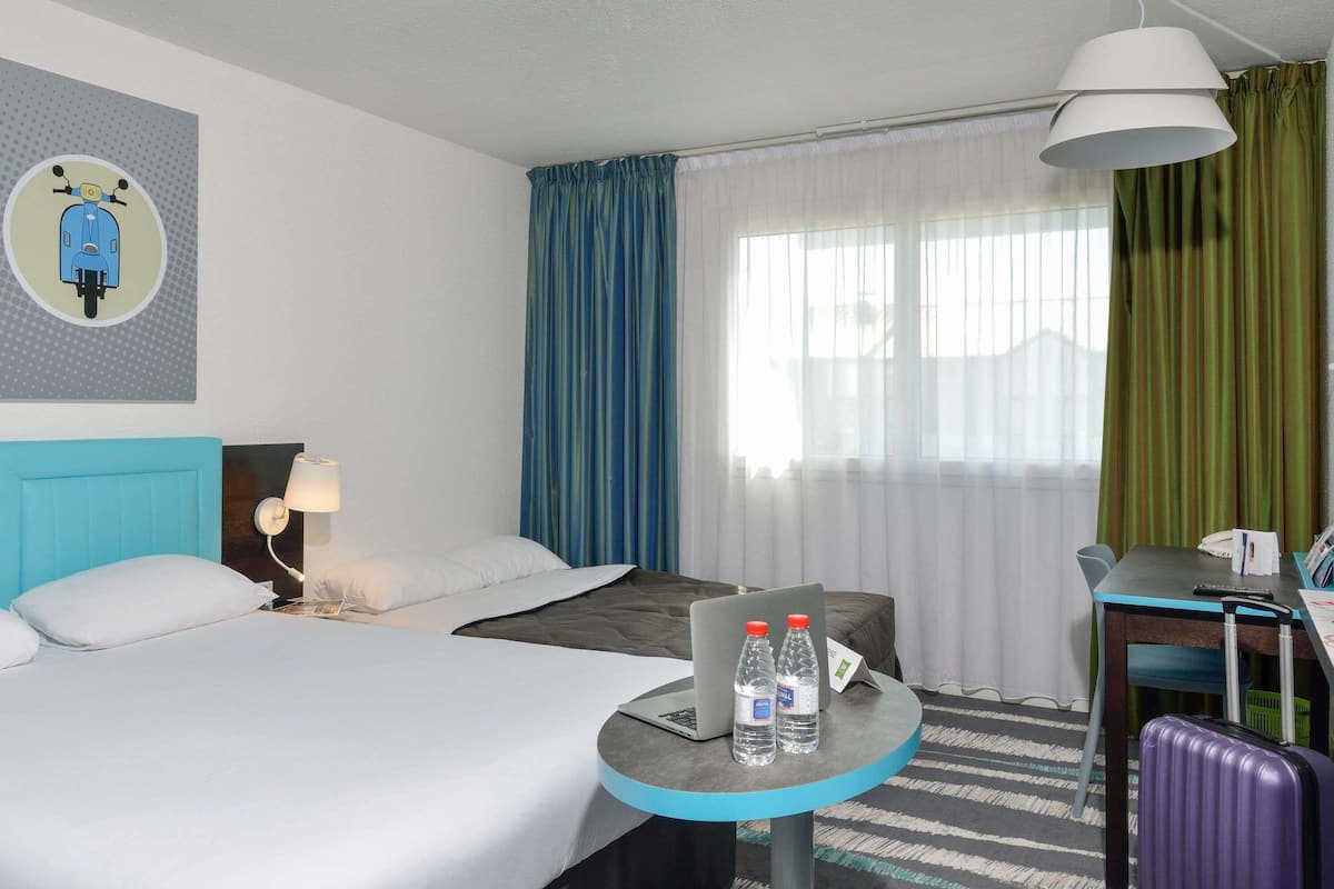 A hotel room with two beds, a desk, a chair, a lamp, a window with curtains, and a wall decoration.
