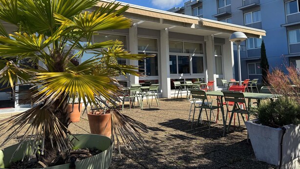 An outdoor seating area with tables and chairs, a palm tree, and a building with multiple windows in the background. - ibis Styles Angouleme Nord (Champniers)