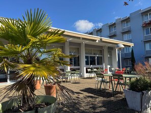Un área de estar al aire libre con mesas y sillas, una palmera y un edificio con varias ventanas al fondo.